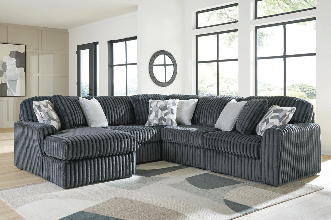 Dark gray L-shaped sectional sofa upholstered in a ribbed plush fabric, featuring deep seats and several throw pillows in solid light gray and a gray/white geometric pattern. The sofa is placed in a modern living room with large windows and a geometric area rug.