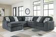 Dark gray L-shaped sectional sofa upholstered in a ribbed plush fabric, featuring deep seats and several throw pillows in solid light gray and a gray/white geometric pattern. The sofa is placed in a modern living room with large windows and a geometric area rug.