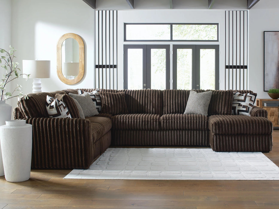 Brown corduroy-textured L-shaped sectional sofa with plush cushions and various throw pillows, including geometric patterns, in a modern living room. The room has light wood floors, a white textured rug, and large windows.