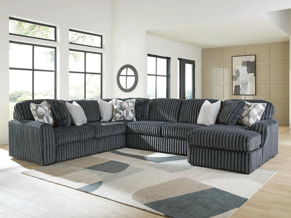 Large U-shaped dark gray sectional sofa with plush ribbed corduroy-like fabric and assorted throw pillows, including patterned and solid gray, in a modern living room with large windows and a geometric rug.