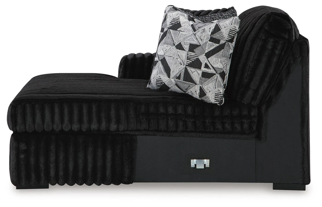 Black faux fur chaise lounge with a ribbed texture, featuring a high right armrest and back. It includes a decorative throw pillow with a black, white, and gray geometric pattern. The base is smooth black with a silver-toned accent.