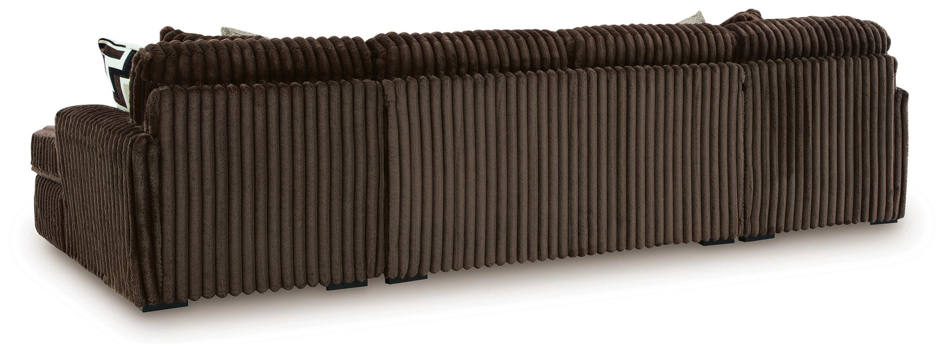 Rear view of a dark brown plush ribbed corduroy sofa or sectional, showing the back and one armrest, with black block feet visible underneath. A patterned throw pillow peeks out from behind the back cushions.