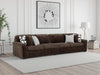 Brown channel-tufted sofa with a corduroy-style texture, featuring four throw pillows (two patterned, two light gray), placed on a light gray rug in a modern living room with white paneled walls.
