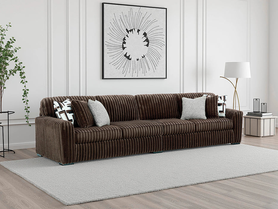 Brown channel-tufted sofa with a corduroy-style texture, featuring four throw pillows (two patterned, two light gray), placed on a light gray rug in a modern living room with white paneled walls.