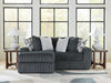 Dark gray plush ribbed fabric L-shaped sectional sofa with light gray and geometric patterned throw pillows, flanked by two wooden end tables with white lamps, on a light wood floor with a geometric rug, in front of large windows.