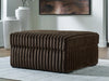 Rectangular brown faux fur ottoman with vertical channel tufting, sitting on a light gray patterned rug in a modern room with white walls, decorative vases, and abstract art.