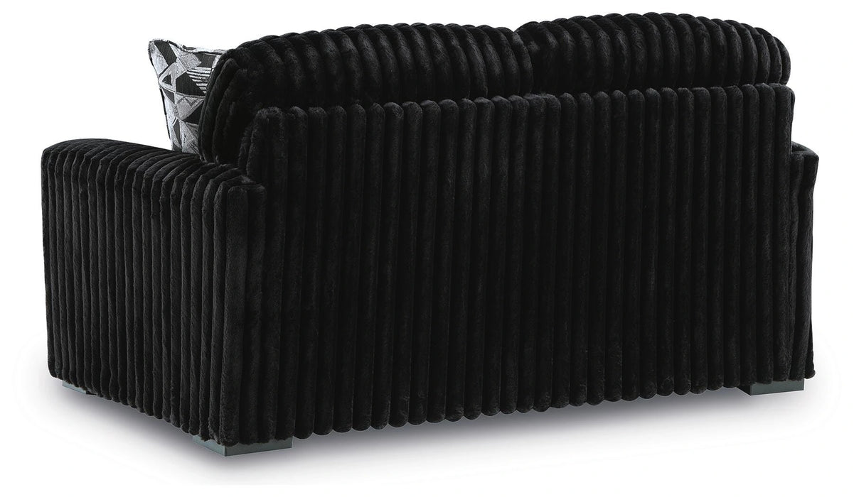 Black channel-tufted loveseat with a plush, ribbed velvet texture. It includes a black and white geometric patterned throw pillow and rests on small dark block feet.