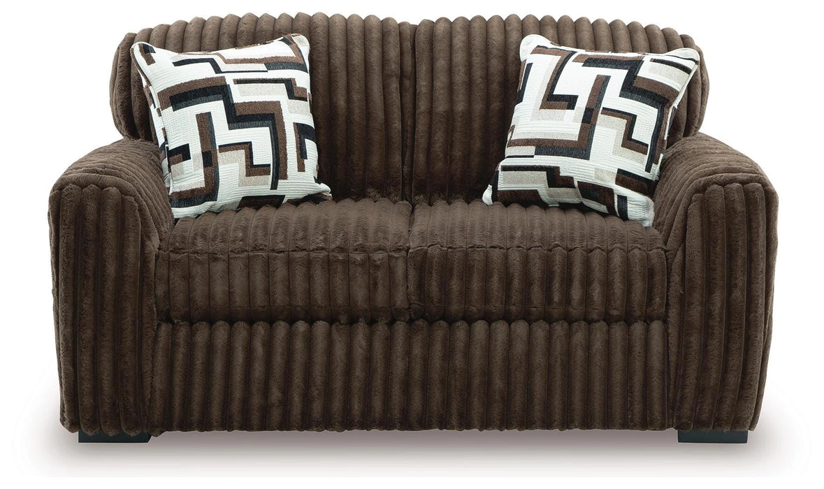 Brown two-seater sofa upholstered in a plush, ribbed fabric. It features two square throw pillows with a modern geometric pattern in white, black, brown, and gray. The sofa has a cozy, contemporary style and sits on dark, low-profile feet.