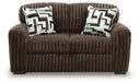 Brown two-seater sofa upholstered in a plush, ribbed fabric. It features two square throw pillows with a modern geometric pattern in white, black, brown, and gray. The sofa has a cozy, contemporary style and sits on dark, low-profile feet.