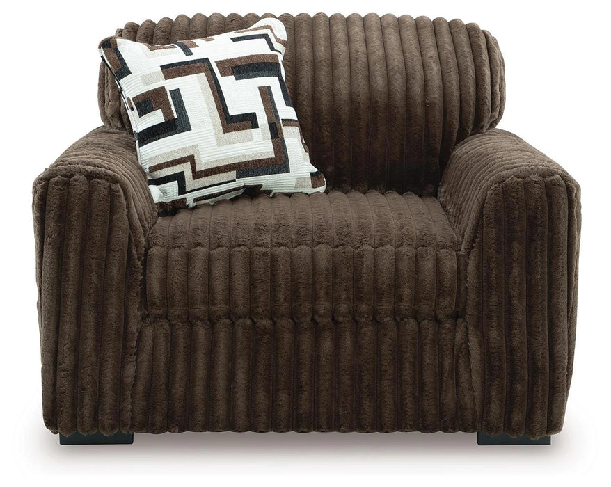 Dark brown plush armchair with a ribbed texture, wide arms, and a low profile. A decorative throw pillow with a white, brown, black, and grey geometric pattern rests on the backrest.