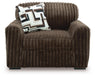 Dark brown plush armchair with a ribbed texture, wide arms, and a low profile. A decorative throw pillow with a white, brown, black, and grey geometric pattern rests on the backrest.