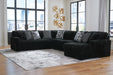 Black L-shaped sectional sofa with a ribbed texture, featuring several geometric and speckled throw pillows. The sofa sits on a light-colored rug over hardwood floors in a modern room with large windows overlooking a city.