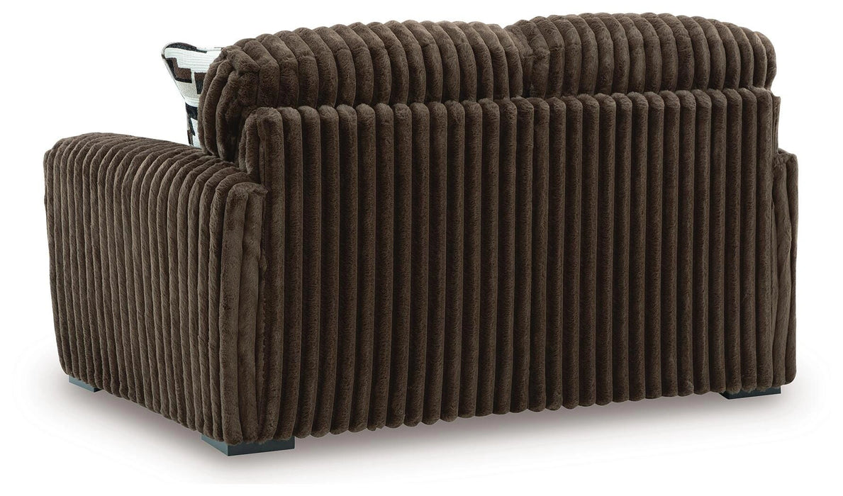 Brown plush ribbed armchair with block feet, seen from the back-right angle. A patterned cushion is partially visible behind the backrest.