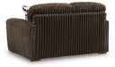 Brown plush ribbed armchair with block feet, seen from the back-right angle. A patterned cushion is partially visible behind the backrest.
