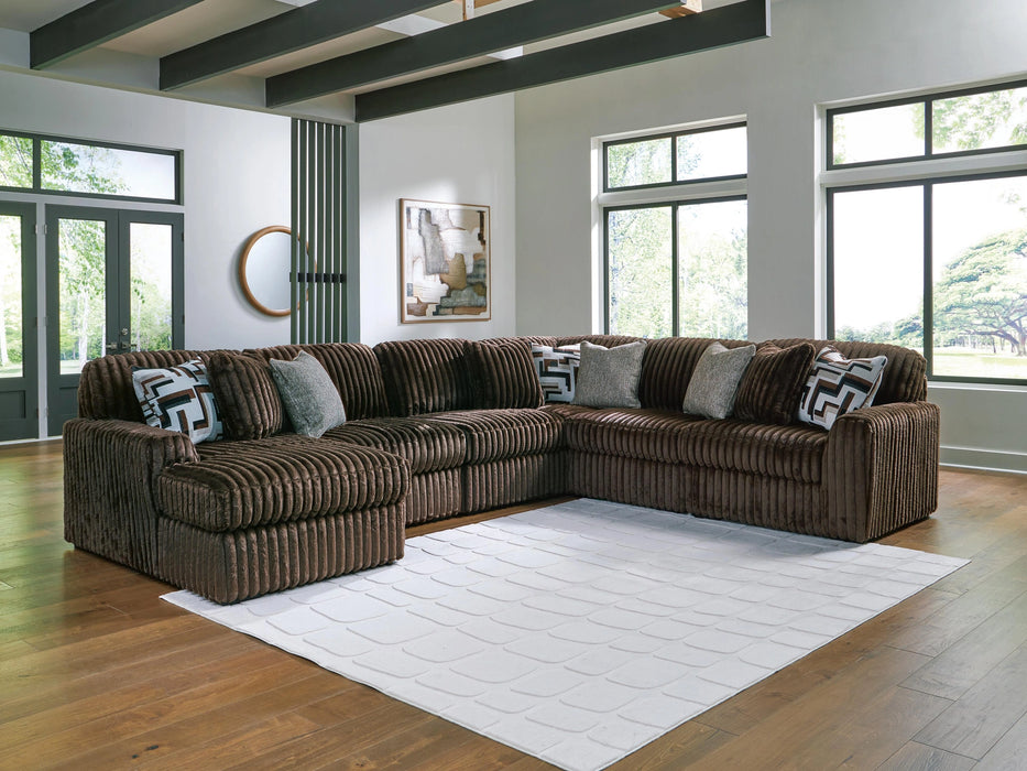 Large brown U-shaped sectional sofa with ribbed fabric texture and plush cushions, accented with patterned and solid grey throw pillows, in a modern living room with hardwood floors and a white textured rug.