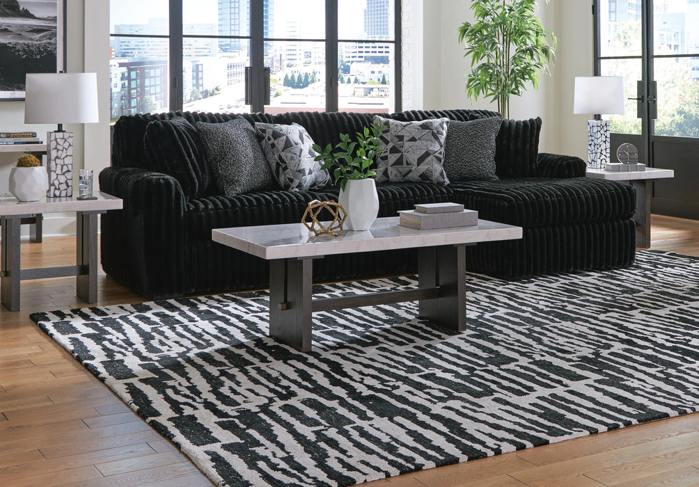 A modern living room features a black channel-tufted sectional sofa with patterned gray and black accent pillows. A rectangular coffee table with a faux marble top and dark wood base sits on an abstract black and white area rug, flanked by matching end tables with white cylindrical lamps and metallic decor.
