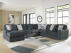 Large U-shaped dark gray sectional sofa with a plush, ribbed corduroy-like texture, featuring several light gray and geometric patterned throw pillows. It sits on a light wood floor with an abstract rug in a bright room with large windows.