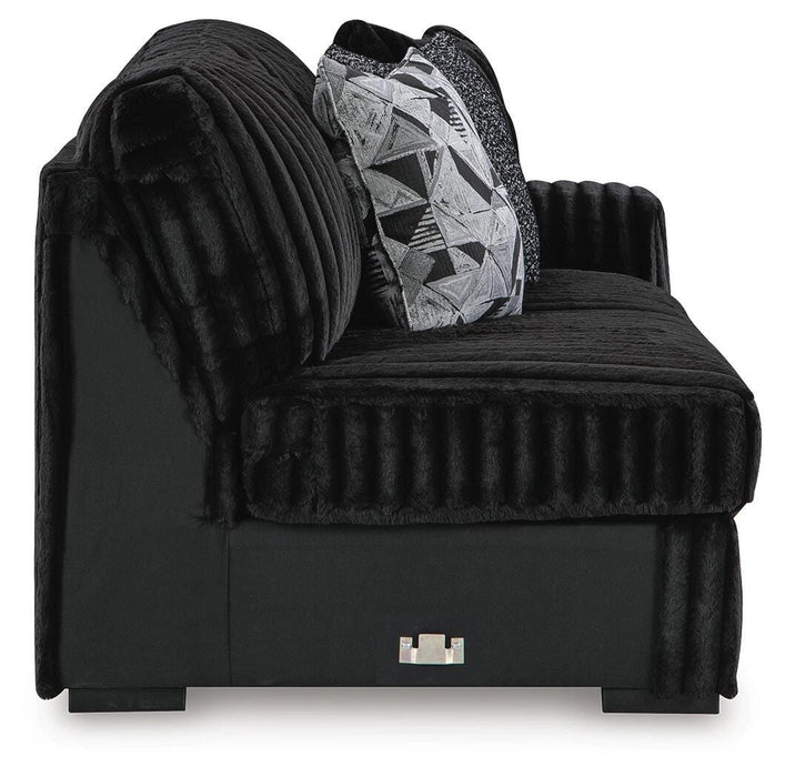 Black modular chair with plush, ribbed faux fur upholstery on cushions and armrest. Features a smooth black fabric base, one geometric patterned throw pillow, and small silver accent on the front.
