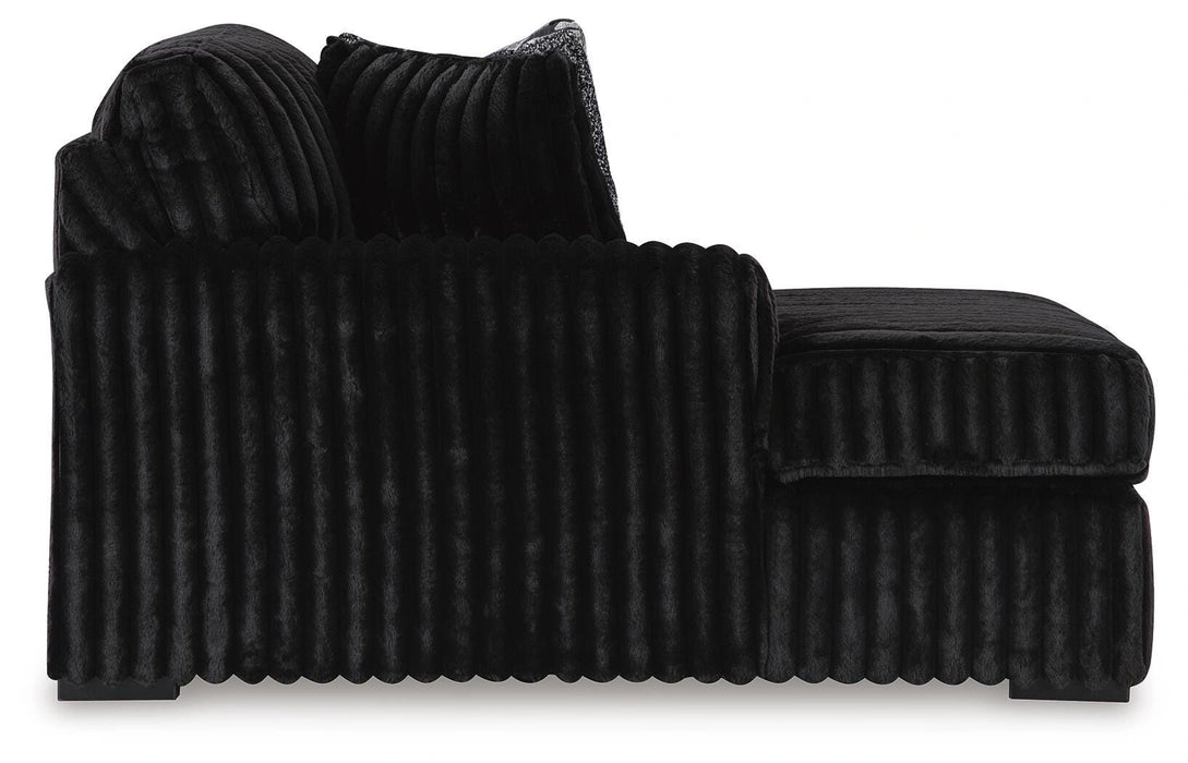 Black chaise lounge upholstered in a soft, channel-tufted plush fabric. It features a single armrest, a deep seat, and a matching back cushion.