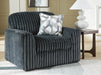 A dark gray plush armchair with a ribbed texture sits on a light geometric rug in a modern living room. A white and gray patterned throw pillow rests on the chair. To the right, a dark wood side table holds a white lamp with a textured base.