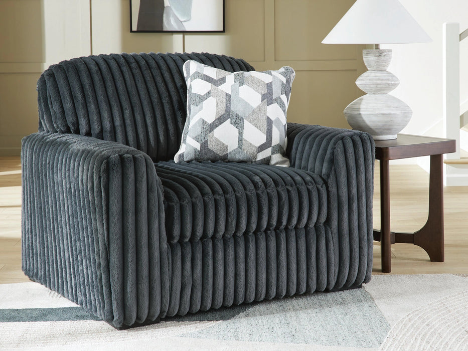 A dark gray plush armchair with a ribbed texture sits on a light geometric rug in a modern living room. A white and gray patterned throw pillow rests on the chair. To the right, a dark wood side table holds a white lamp with a textured base.