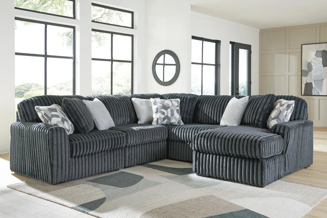 Dark gray L-shaped sectional sofa with plush, ribbed corduroy-like fabric, featuring several light gray and geometric patterned throw pillows. The sofa is placed on a light-colored rug in a bright room with large windows.