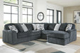Dark gray L-shaped sectional sofa with plush, ribbed corduroy-like fabric, featuring several light gray and geometric patterned throw pillows. The sofa is placed on a light-colored rug in a bright room with large windows.
