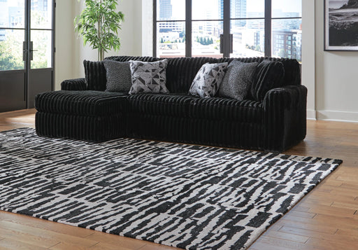 A black ribbed sectional sofa with four decorative throw pillows sits on a black and white abstract patterned area rug. The room features light hardwood floors, light walls, and large windows overlooking a city skyline.