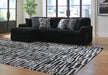 A black ribbed sectional sofa with four decorative throw pillows sits on a black and white abstract patterned area rug. The room features light hardwood floors, light walls, and large windows overlooking a city skyline.