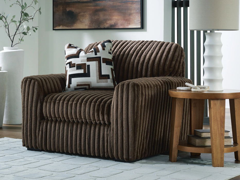 A brown armchair with a plush, ribbed texture, featuring a decorative throw pillow with a geometric pattern in brown, black, and white. The chair is placed on a light-colored rug next to a wooden side table with a white lamp.