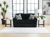 Black loveseat with a ribbed, textured fabric, featuring two geometric black and white throw pillows. It sits on a light wood floor with a white striped rug, flanked by marble-top end tables with lamps, in a modern room with large windows overlooking a city.