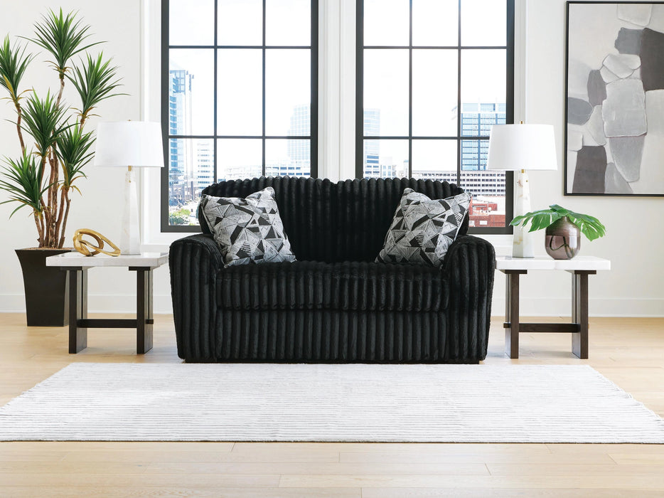 Black loveseat with a ribbed, textured fabric, featuring two geometric black and white throw pillows. It sits on a light wood floor with a white striped rug, flanked by marble-top end tables with lamps, in a modern room with large windows overlooking a city.