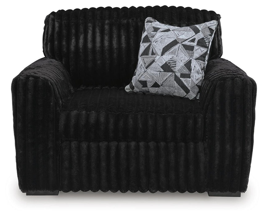 Black plush armchair with a ribbed texture, featuring rounded arms and back. A decorative throw pillow with a black, white, and gray geometric pattern rests on the seat.
