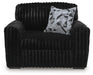 Black plush armchair with a ribbed texture, featuring rounded arms and back. A decorative throw pillow with a black, white, and gray geometric pattern rests on the seat.