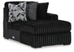 Black plush ribbed fabric chaise lounge with a right-side armrest and black block feet. It features two decorative pillows: one with a black, white, and gray geometric pattern, and another solid gray textured pillow.