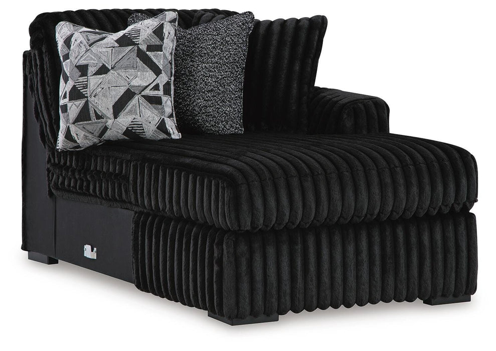 Black plush ribbed fabric chaise lounge with a right-side armrest and black block feet. It features two decorative pillows: one with a black, white, and gray geometric pattern, and another solid gray textured pillow.