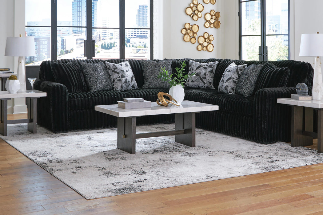 Modern living room with a large black ribbed sectional sofa, accented with grey patterned pillows. A white marble top coffee table and two matching end tables sit on a light grey and black abstract rug. White marble lamps and gold wall decor complete the space.