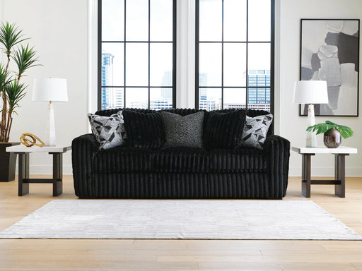 Black ribbed sofa with three decorative pillows (two geometric, one textured black) in a modern living room. It sits on a white striped rug, flanked by white marble-top side tables with lamps. Large windows show a city view, and a gray abstract painting hangs on the wall.