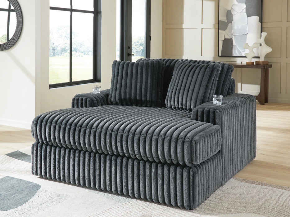 Oversized dark gray chair with plush, ribbed fabric, deep seating, and wide arms. It features two matching back cushions and sits on a light-colored rug in a bright room with large windows.