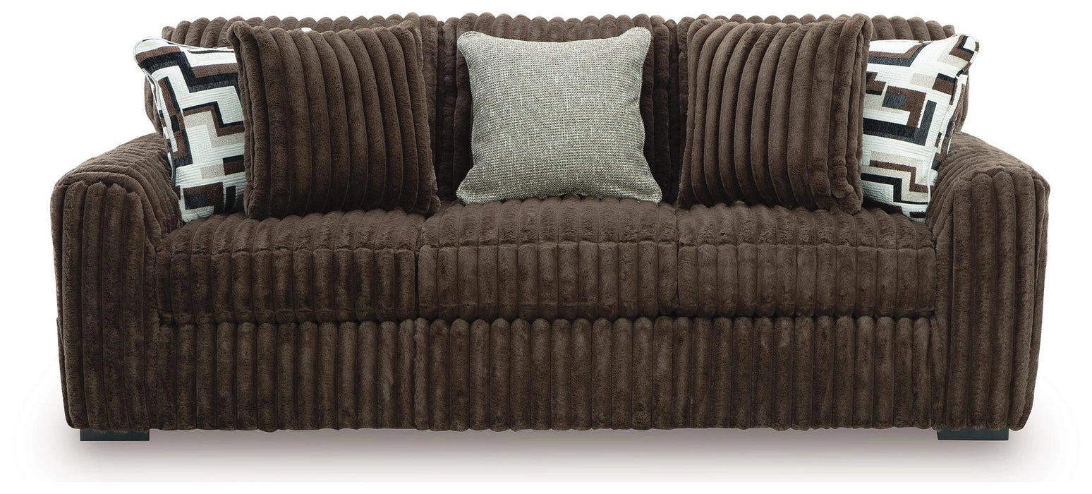 Brown ribbed chenille sofa with three matching back cushions. It includes two throw pillows with a geometric pattern in brown, white, black, and gray, and one light gray textured throw pillow.