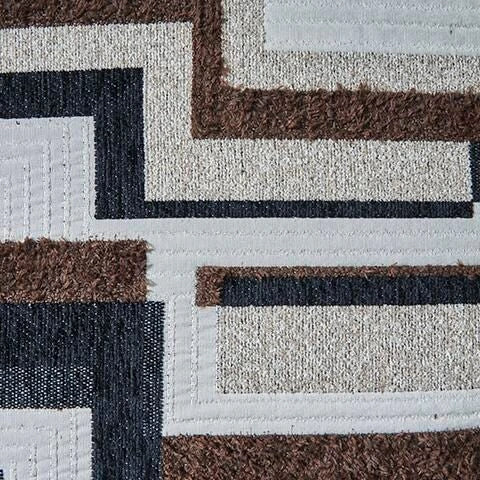 Close-up of a fabric swatch featuring a geometric pattern in brown, black, beige, and white. The pattern combines flat woven, shaggy, and quilted textures.