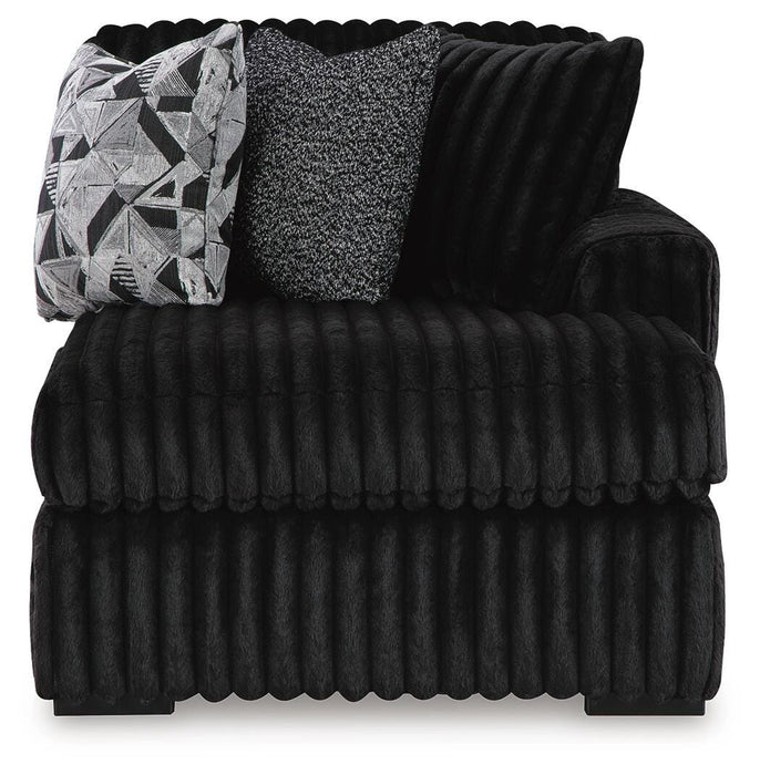 Oversized black ribbed plush armchair featuring three decorative pillows: one matching ribbed black, one black and white speckled, and one black and white geometric pattern.