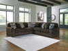 Dark brown L-shaped sectional sofa with plush, ribbed corduroy-like fabric, featuring several throw pillows in patterned and textured gray designs. The sofa sits on a wood floor with a white geometric rug in a bright, modern living room with large windows.