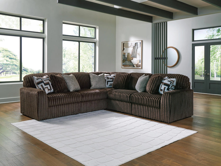 Dark brown L-shaped sectional sofa with plush, ribbed corduroy-like fabric, featuring several throw pillows in patterned and textured gray designs. The sofa sits on a wood floor with a white geometric rug in a bright, modern living room with large windows.