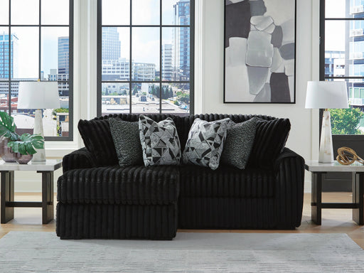 Black ribbed sectional sofa with patterned and textured throw pillows in a modern living room with large windows overlooking a city, light rug, and marble-top side tables with lamps.