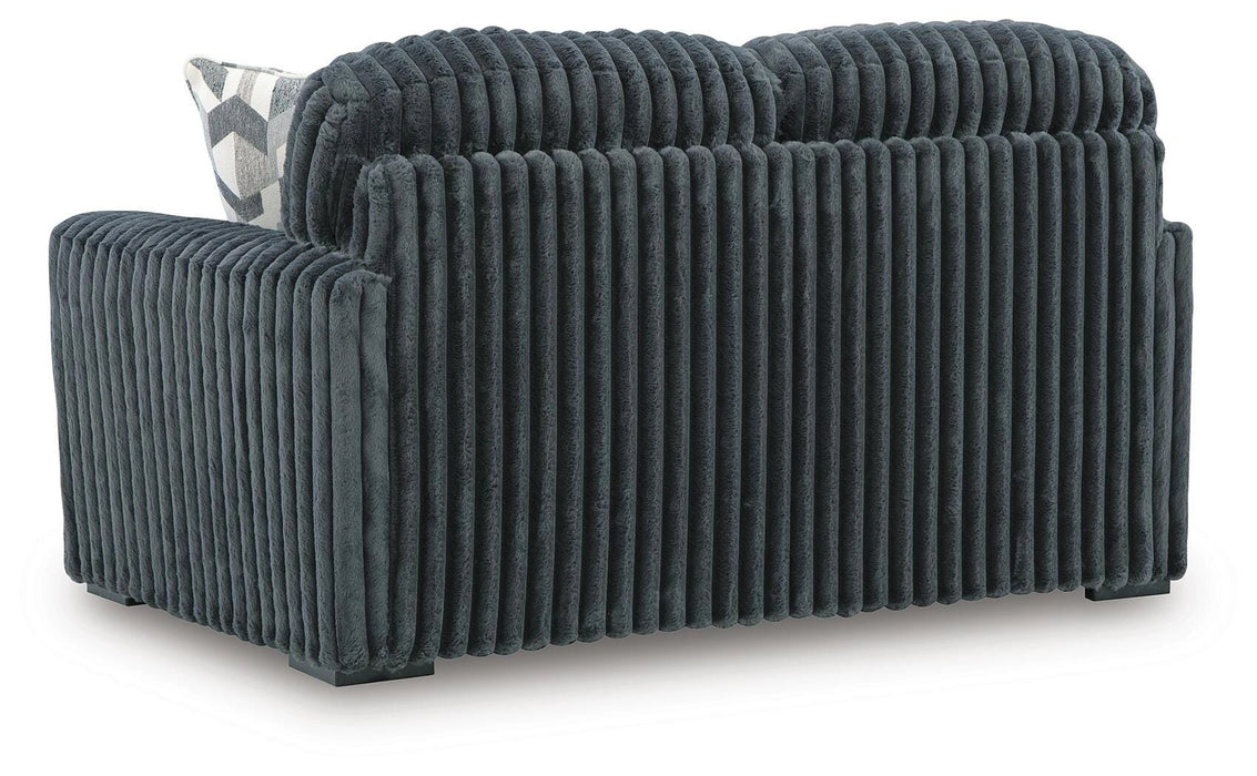 Dark gray ribbed sofa or loveseat, upholstered in a plush, corduroy-like fabric. The back and side are visible, showing vertical ribs. A white and gray geometric patterned throw pillow rests on the left armrest. It sits on dark, low-profile block feet.