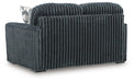 Dark gray ribbed sofa or loveseat, upholstered in a plush, corduroy-like fabric. The back and side are visible, showing vertical ribs. A white and gray geometric patterned throw pillow rests on the left armrest. It sits on dark, low-profile block feet.