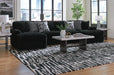 Black ribbed sectional sofa with throw pillows in a modern living room. The sofa is paired with a marble-top coffee table, end tables with lamps, and a black and white abstract patterned rug on a hardwood floor. Large windows overlook a city skyline.