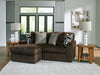 Dark brown ribbed plush sectional sofa with an attached ottoman, accented with patterned and textured throw pillows, in a bright living room with large windows, wooden side tables, and white lamps.