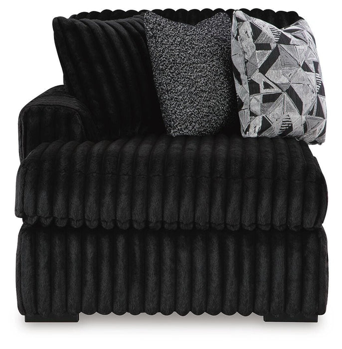 Black plush armchair with a ribbed texture, featuring a wide seat and back. It includes two accent pillows: one speckled gray and one with a black, white, and gray geometric pattern. The chair has low, dark feet.